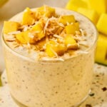 A jar of mango overnight oats topped with fresh diced mango and shredded coconut. Behind the jar is a cut mango.