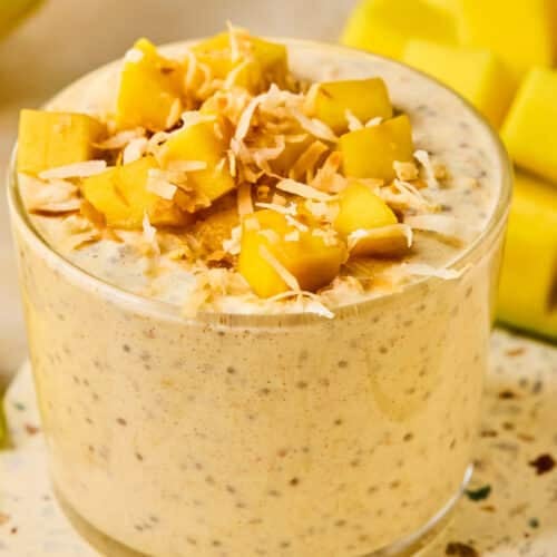 A jar of mango overnight oats topped with fresh diced mango and shredded coconut. Behind the jar is a cut mango.