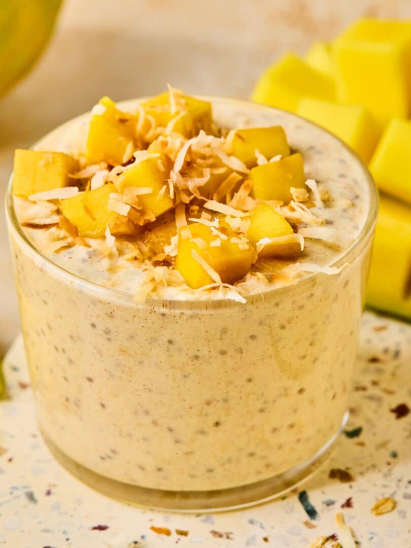 A jar of mango overnight oats topped with fresh diced mango and shredded coconut. Behind the jar is a cut mango.