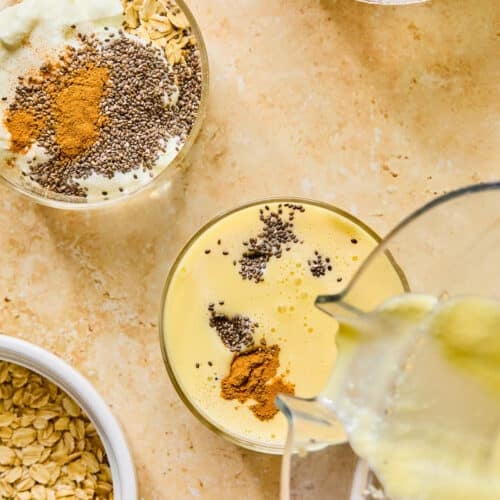Overhead shot of a glass jar filled with rolled oats, greek yogurt, chia seeds, and cinnamon, with mango milk being poured into it. Next to the jar is another jar with more oats, yogurt, chia seeds, and cinnamon, a bowl of chia seeds, a bowl of overnight oats, and a bowl of greek yogurt.