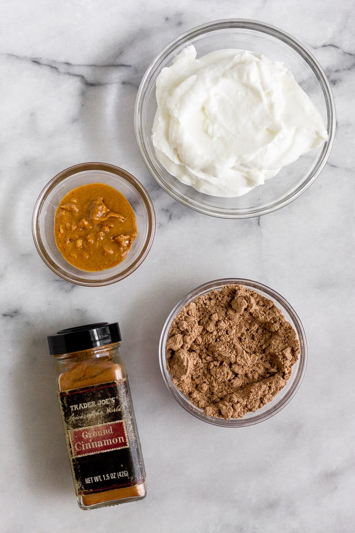 Encimera blanca con un bol de yogur griego, un bol de proteína de chocolate en polvo, un bol de mantequilla de cacahuete y un bote de canela.