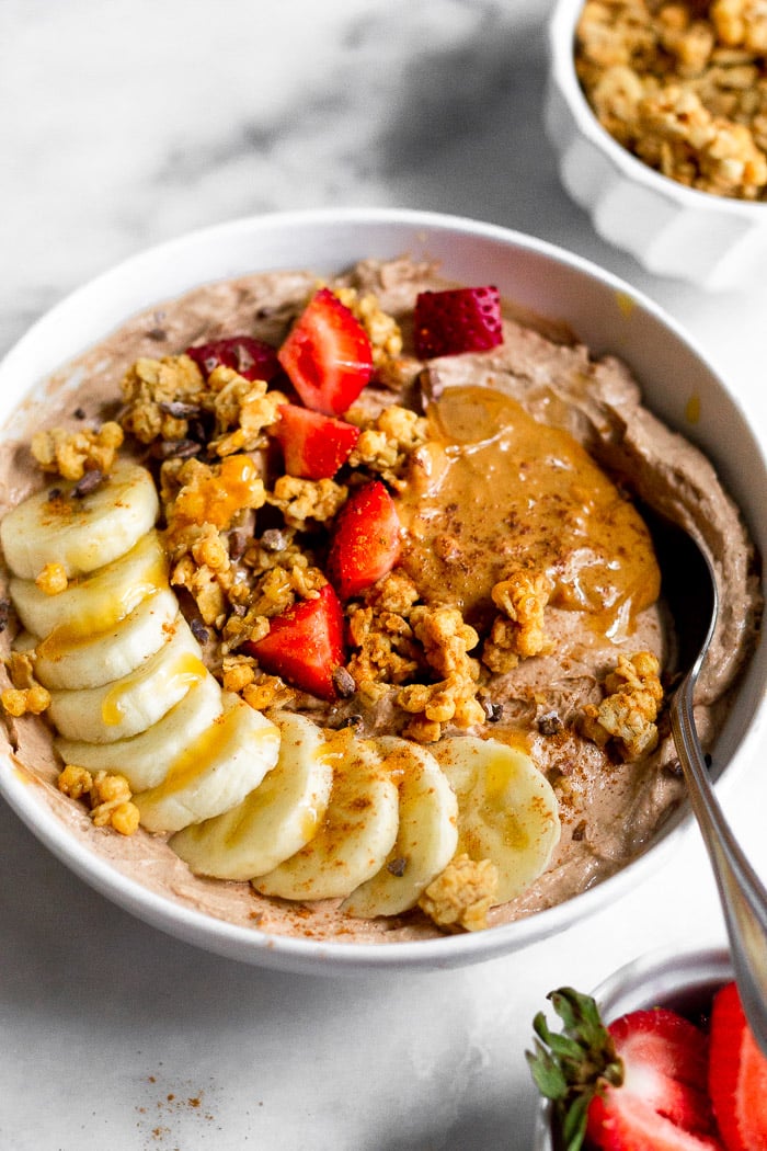 Tazón de yogur proteico de chocolate y mantequilla de cacahuete cubierto con plátano, granola, fresas, mantequilla de cacahuete y miel con una cuchara saliendo de él. Delante hay un pequeño bol de fresas y detrás un pequeño bol de granola.