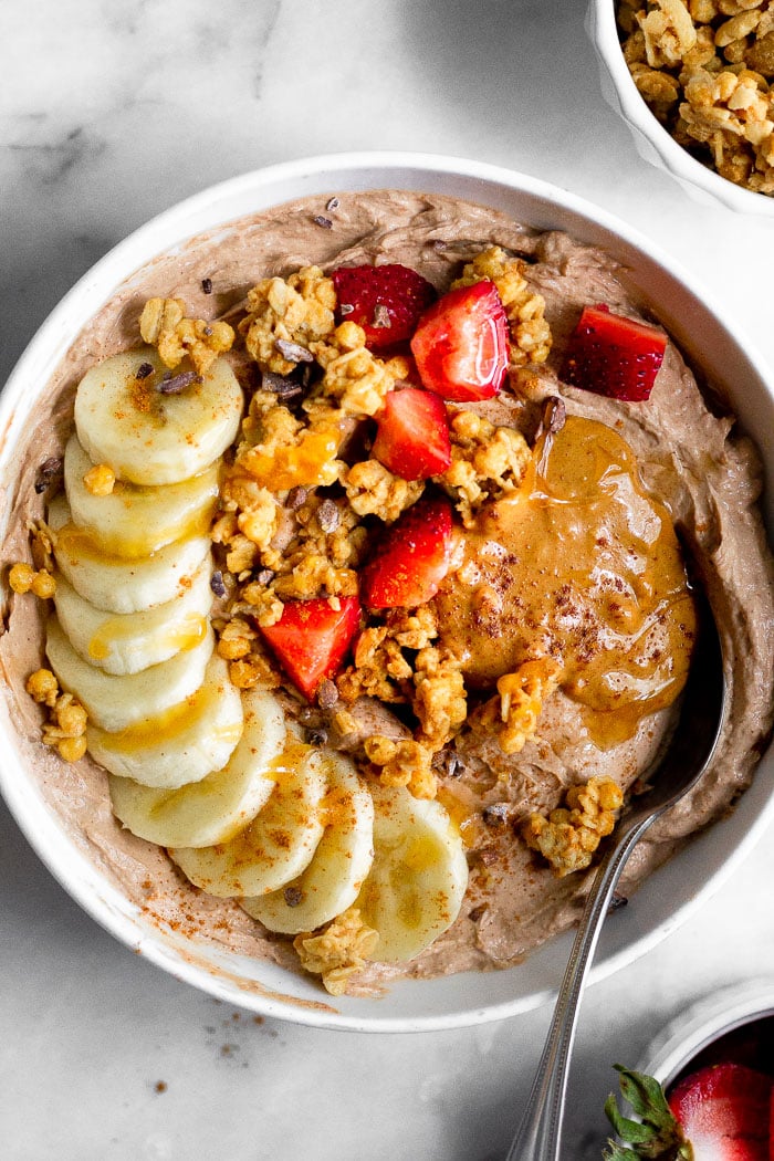 Tazón de yogur proteico con plátano, granola, fresas, mantequilla de cacahuete y miel. Una cuchara sale del bol.