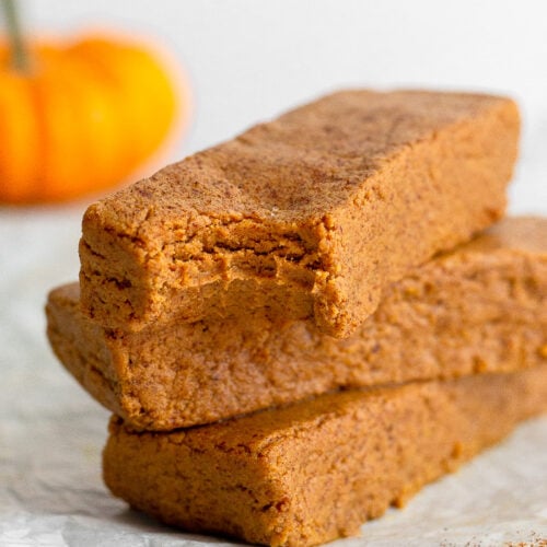 A stack of 3 pumpkin protein bars with a bite taken out of the first one. In the background is a small pumpkin.