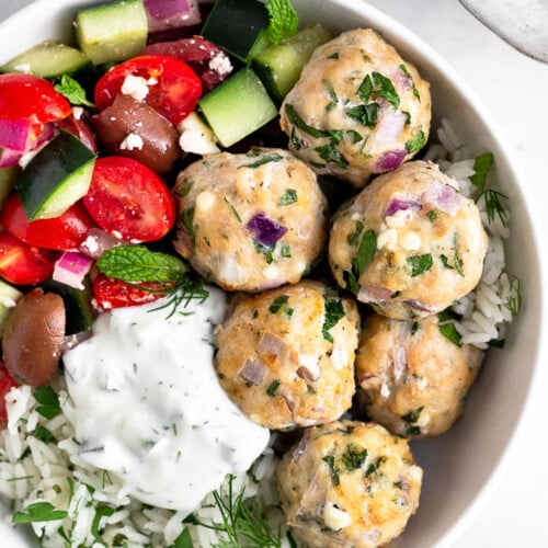 Greek chicken meatballs with herby rice, a greek salad with tomatoes, cucumbers, olives, and feta, and tzatziki sauce in a white bowl.