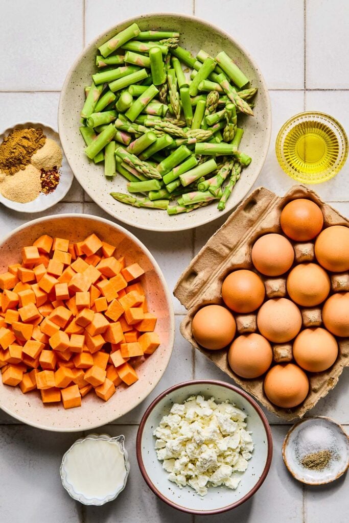 A large bowl of chopped asparagus, a small bowl of olive oil, a carton of a dozen eggs, a small plate with salt and pepper, a bowl with crumbled goat cheese, a cup of milk, a bowl of raw diced sweet potatoes, and a plate with a few different piles of spices on it on a white tile counter.