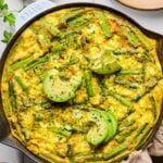 Asparagus frittata with sweet potatoes and goat cheese in a large enameled skillet. It is topped with sliced avocado and fresh parsley. Around the pan is a tan kitchen towel, a plate with two forks on it, a small dish of chopped fresh parsley, and another empty plate.