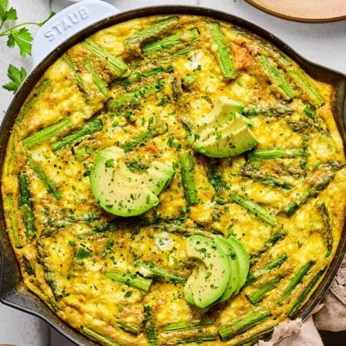 Asparagus frittata with sweet potatoes and goat cheese in a large enameled skillet. It is topped with sliced avocado and fresh parsley. Around the pan is a tan kitchen towel, a plate with two forks on it, a small dish of chopped fresh parsley, and another empty plate.