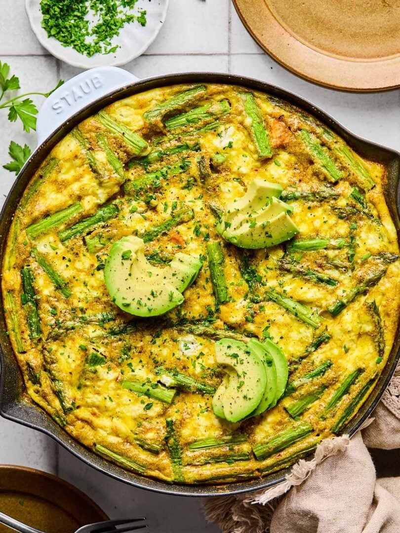Asparagus frittata with sweet potatoes and goat cheese in a large enameled skillet. It is topped with sliced avocado and fresh parsley. Around the pan is a tan kitchen towel, a plate with two forks on it, a small dish of chopped fresh parsley, and another empty plate.