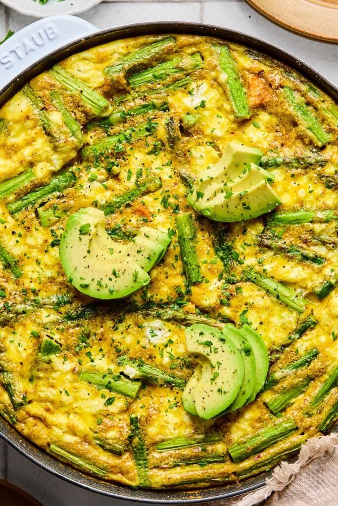 Close up overhead shot of a frittata with asparagus, sweet potatoes, and goat cheese in a large skillet. It is topped with sliced avocado and fresh chopped parsley.