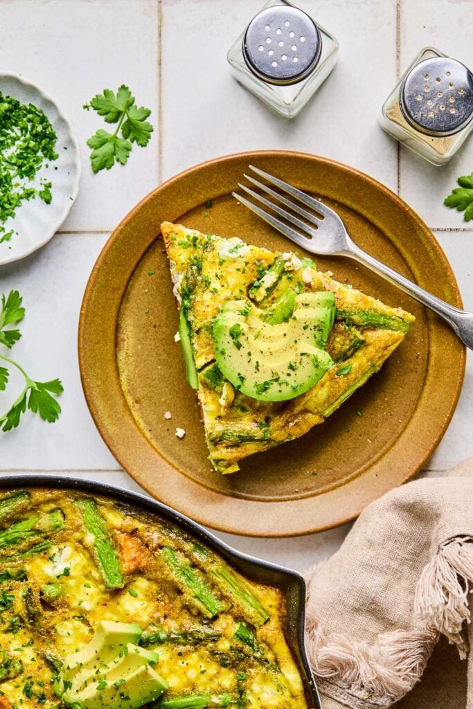 A piece of asparagus and goat cheese frittata and a metal fork on a brown plate. The frittata is topped with avocado and fresh parsley. Next to the plate is a tan kitchen towel, a large skillet with the fresh of the frittata in it, a plate with fresh parsley, and a salt and pepper shaker.