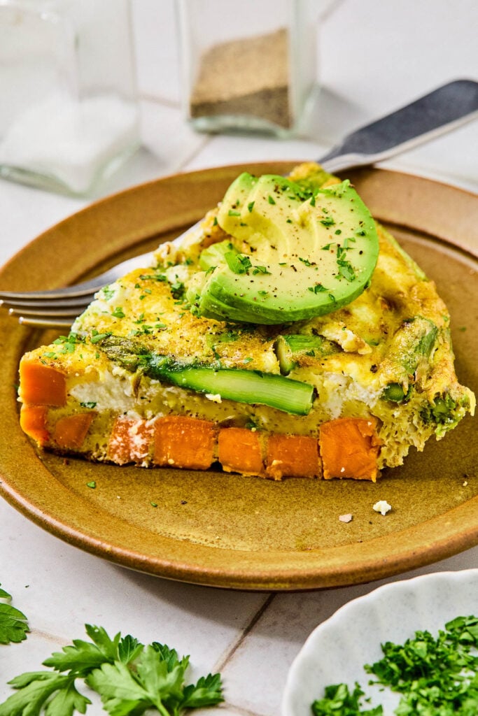 A close up of a plate with a piece of asparagus and sweet potato frittata topped with avocado and fresh parsley. There is also a fork on the plate. Around the plate is a plate of chopped parsley, some fresh parsley sprigs, and a salt and pepper shaker.