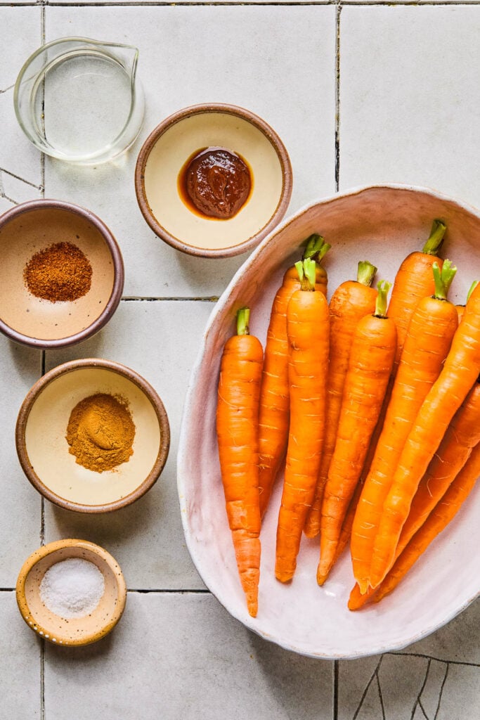 A large oval platter of large peeled carrots, a small bowl of salt, a small bowl of cinnamon, a small bowl of chili powder, a small bowl of chipotle paste, and a small cup of melted coconut oil on a white tile countertop.