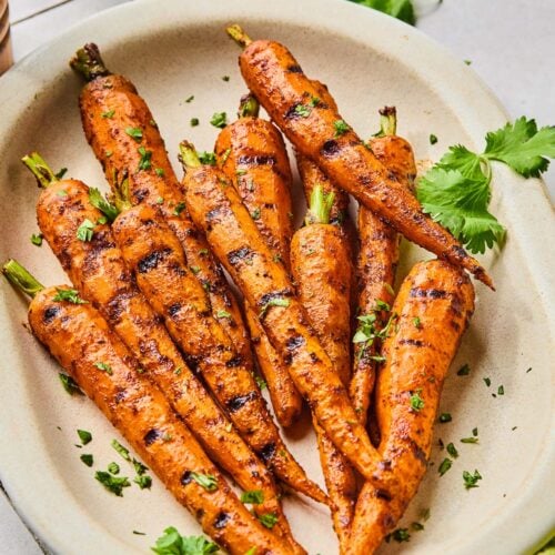 A serving platter of grilled carrots topped with fresh cilantro. Around the platter is a green kitchen towel, more fresh cilantro, and a salt and pepper shaker.