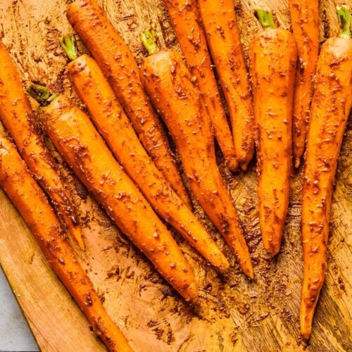 Raw, whole, peeled carrots coated with oil and spices on a wooden cutting board.
