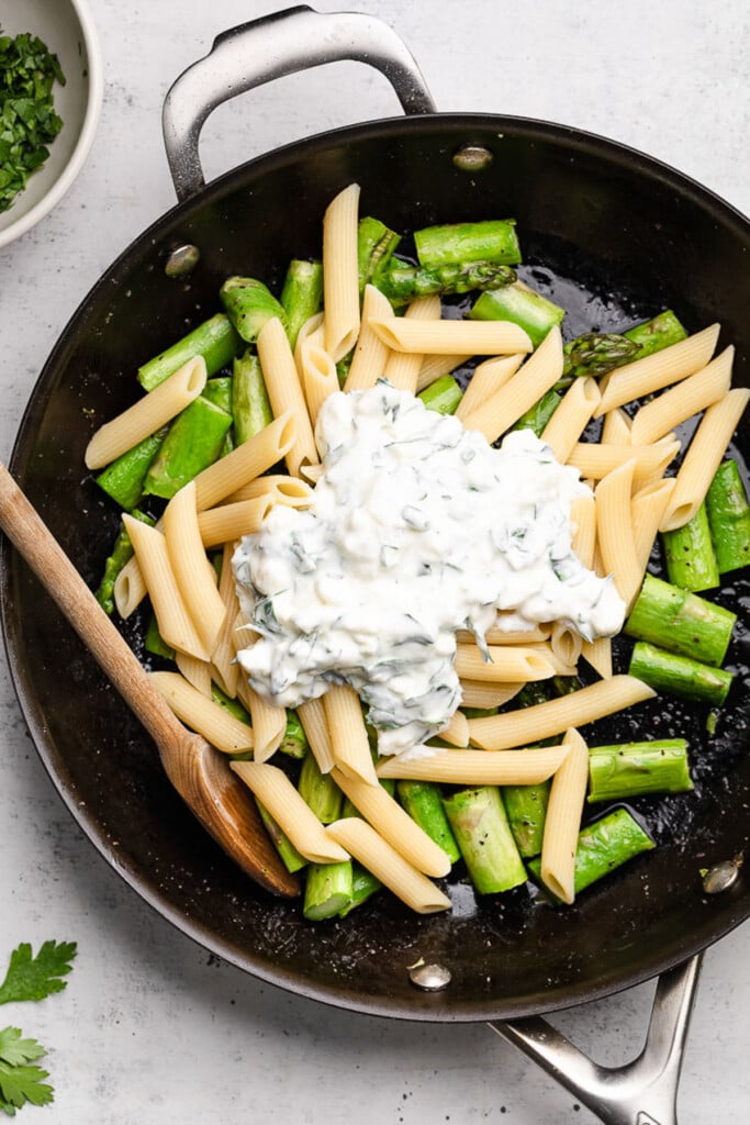 Skillet filled with chopped saut&eacute;ed asparagus, cooked penne, and a thick and creamy white sauce before it is mixed together. A wooden spoon is in the pan as well.