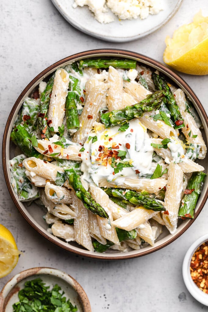 A bowl of lemon asparagus pasta tossed in a creamy yogurt sauce topped with red pepper flakes. Around the bowl is a dish of chopped herbs, some lemons, a bowl of feta, and a small dish of red pepper flakes.