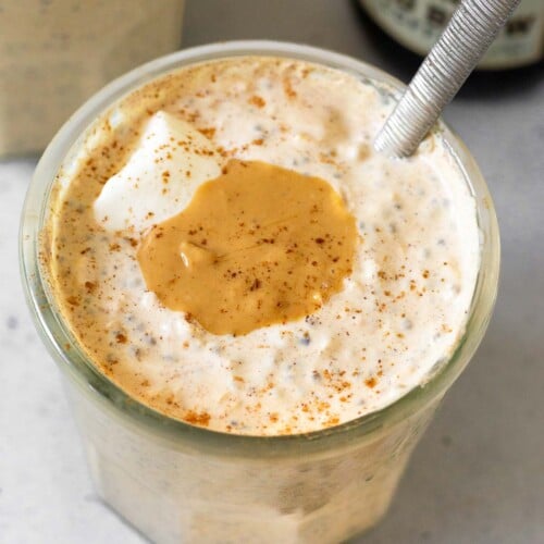 A jar of coffee overnight oats topped with yogurt, cashew butter, and cinnamon with a spoon in the jar. Behind it is another jar of overnight oats and bottle of cold brew coffee. In front of the jar is a teaspoon of cinnamon and some coffee beans.