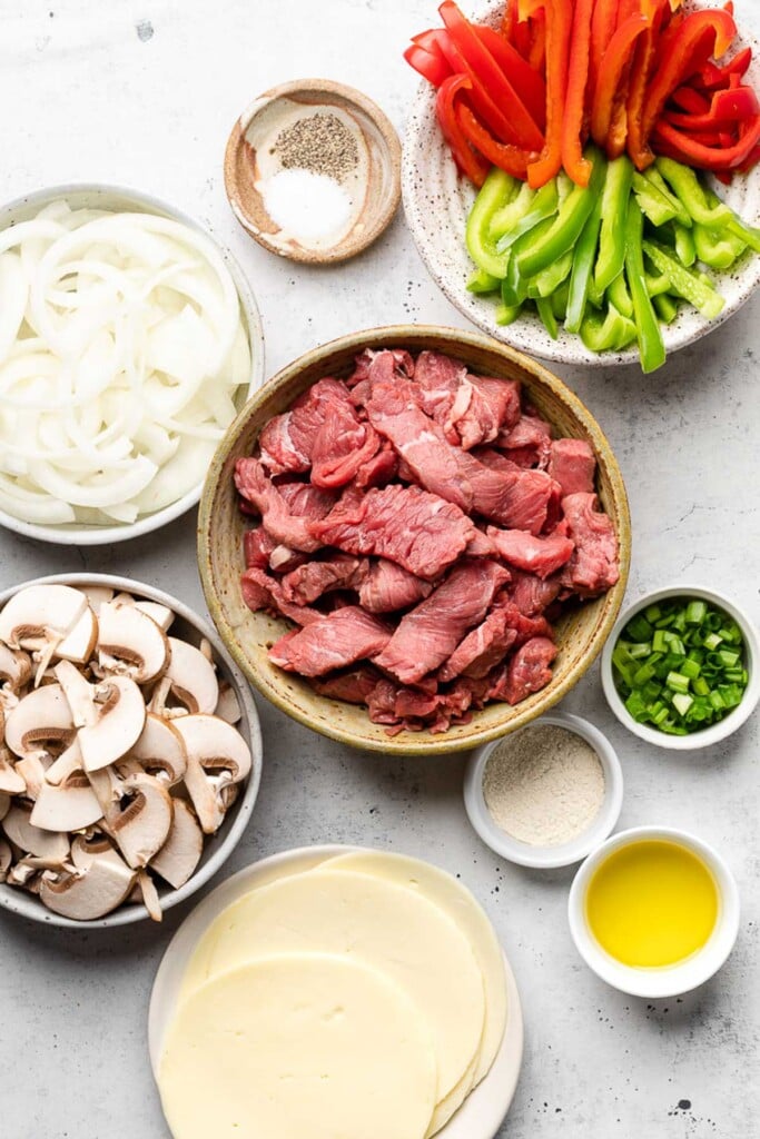 Concrete counter with a bowl of julienned red and green bell peppers, a bowl of thinly sliced steak, a small bowl of sliced green onions, a small bowl of garlic powder, a small bowl of olive oil, a plate of sliced provolone cheese, a bowl of sliced mushrooms, a bowl of sliced onions, and a small bowl of salt and pepper.