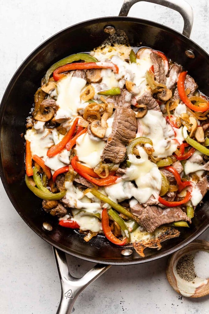 Cheesesteak skillet in a large pan with peppers, onions, and mushrooms topped with melted cheese. Next to the pan is a small bowl of salt and pepper.