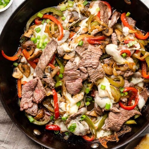 Philly cheesesteak skillet with peppers and onions and topped with melted cheese in a large pan. It is garnished with sliced green onions. Around the pan is a plate of toasted bread, a tan linen towel, and a small bowl of sliced green onions.