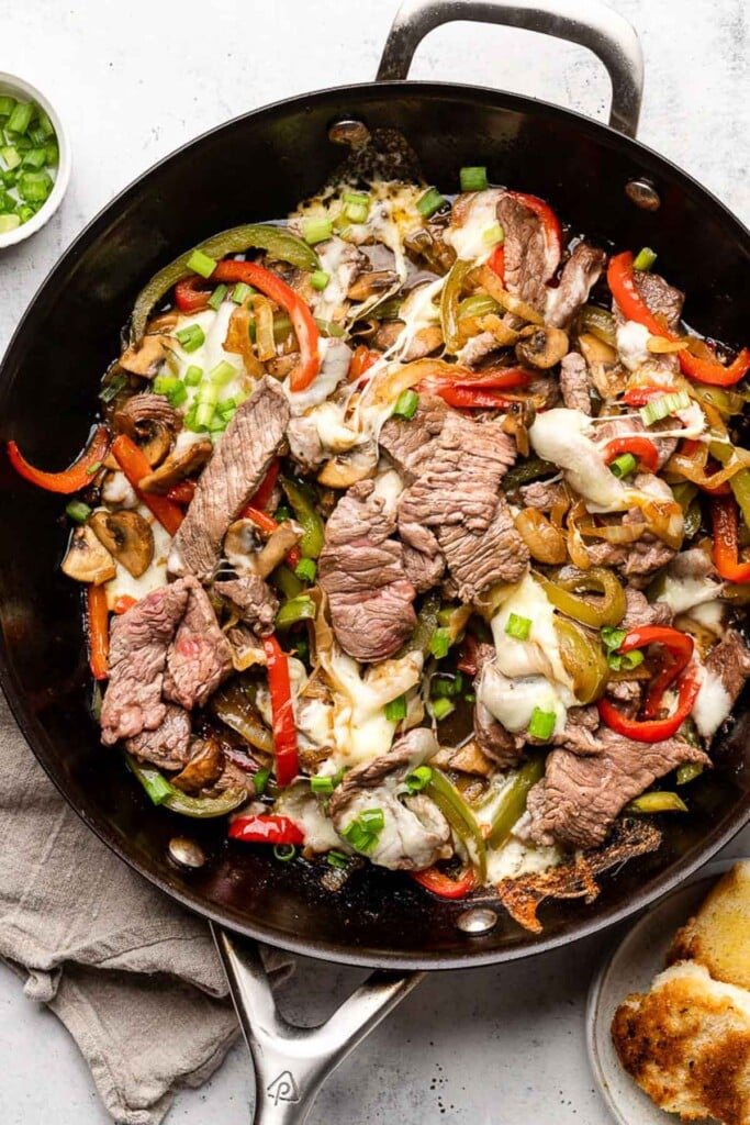 Philly cheesesteak skillet with peppers and onions and topped with melted cheese in a large pan. It is garnished with sliced green onions. Around the pan is a plate of toasted bread, a tan linen towel, and a small bowl of sliced green onions.