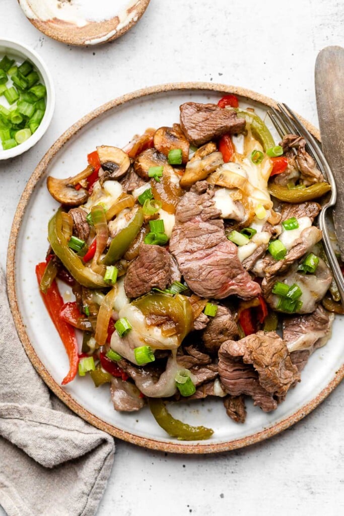 A plate of Philly cheesesteak skillet with peppers, onions, mushrooms, and melted cheese topped with sliced green onions. A fork and a knife is on the plate and around the plate is a tan linen towel and a bowl of sliced green onions.