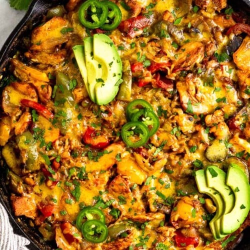 Overhead shot of chicken fajita casserole topped with melted cheese in large cast iron skillet. It is garnished with sliced avocado, sliced jalapeños, and cilantro. Around the pan is striped kitchen towel, a bun of cilantro, and lime wedges.