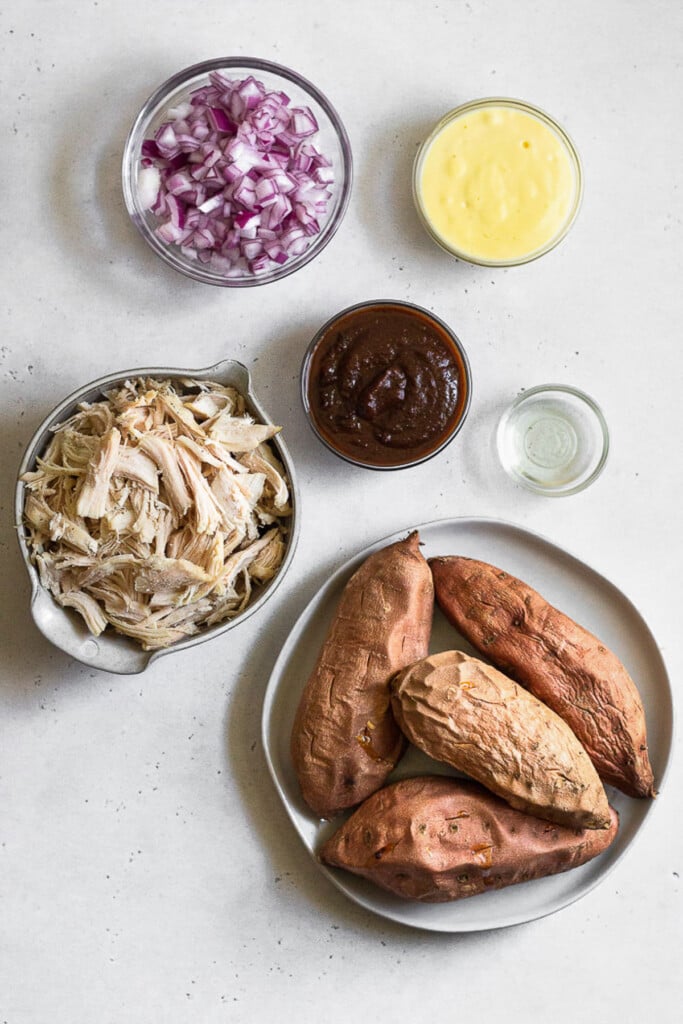 Small bowl of mayonnaise, a bowl of BBQ sauce, a bowl of olive oil, a plate with 4 cooked sweet potatoes on it, a bowl of shredded chicken, and a bowl of diced red onions on a grey concrete counter.