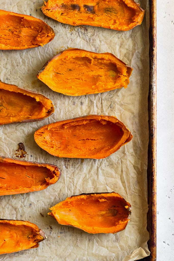 Cooked sweet potato halves on a baking sheet that have the insides scooped out of them and just the skins remain.