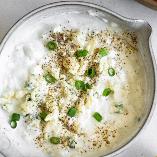 Overhead shot of greek yogurt blue cheese dressing topped with green onions and pepper. Next to it is half a lemon and plate of carrots and celery.