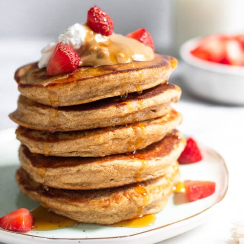 A stack of protein pancakes on a plate topped with yogurt, peanut butter, and strawberries with maple syrup dripping down them. Behind the plate is a bowl of diced strawberries and a jar of milk.