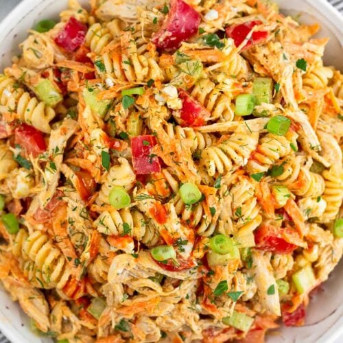 Buffalo chicken pasta salad in a large white bowl. It is on a striped towel and next to it a bowl of herbs.