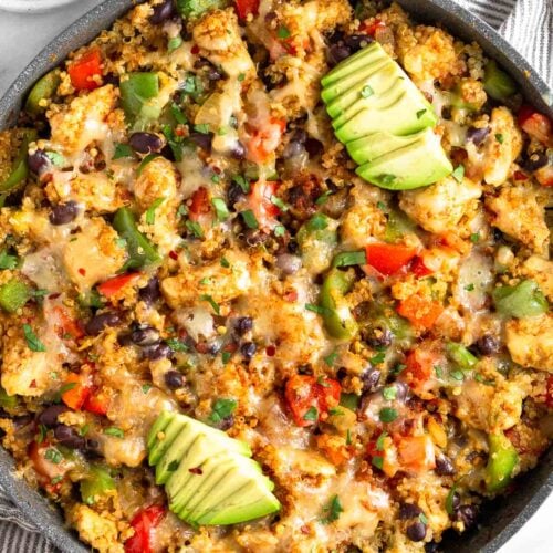 Chicken quinoa skillet in a large pan with veggies and topped with melted cheese topped with avocado. Around the pan is an half of avocado and a bowl of sliced lime.