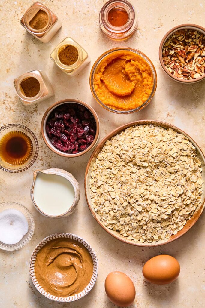 A small jar of maple syrup, a bowl of chopped pecans, a bowl of pumpkin puree, a large bowl of rolled oats, two brown eggs, a bowl of almond butter, a small bowl of salt, a small pitcher of milk, a small bowl of maple syrup, a bowl of dried cranberries, and three different spice jars on a tan concrete counter.