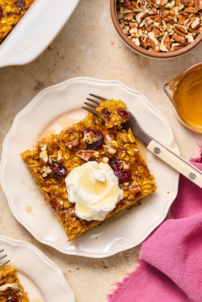 A piece of baked pumpkin oatmeal on a plate topped with greek yogurt and maple syrup. A little piece is taken off the corner and is sitting on a fork on the plate. Around the plate is a pink napkin, another plate with a piece of oatmeal on it, a baking dish with oatmeal, a bowl of chopped pecans, and a small cup of maple syrup.