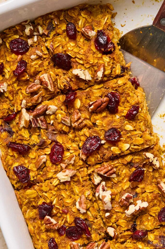 A close up of a baking dish filled with baked pumpkin oatmeal topped with pecans and dried cranberries.