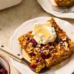 A piece of pumpkin baked oatmeal with cranberries and pecans on a white plate. It is topped with greek yogurt and a drizzle of maple syrup. A little piece is taken off the corner and is sitting on a fork on the plate. Around the plate is a pink napkin, a dish of dried cranberries, a baking dish with baked oatmeal, and another plate with a piece of baked oatmeal.