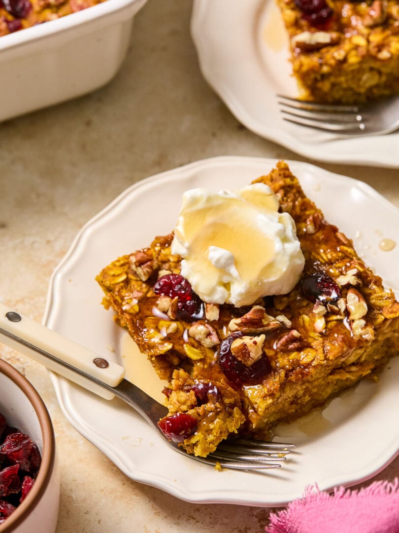 A piece of pumpkin baked oatmeal with cranberries and pecans on a white plate. It is topped with greek yogurt and a drizzle of maple syrup. A little piece is taken off the corner and is sitting on a fork on the plate. Around the plate is a pink napkin, a dish of dried cranberries, a baking dish with baked oatmeal, and another plate with a piece of baked oatmeal.