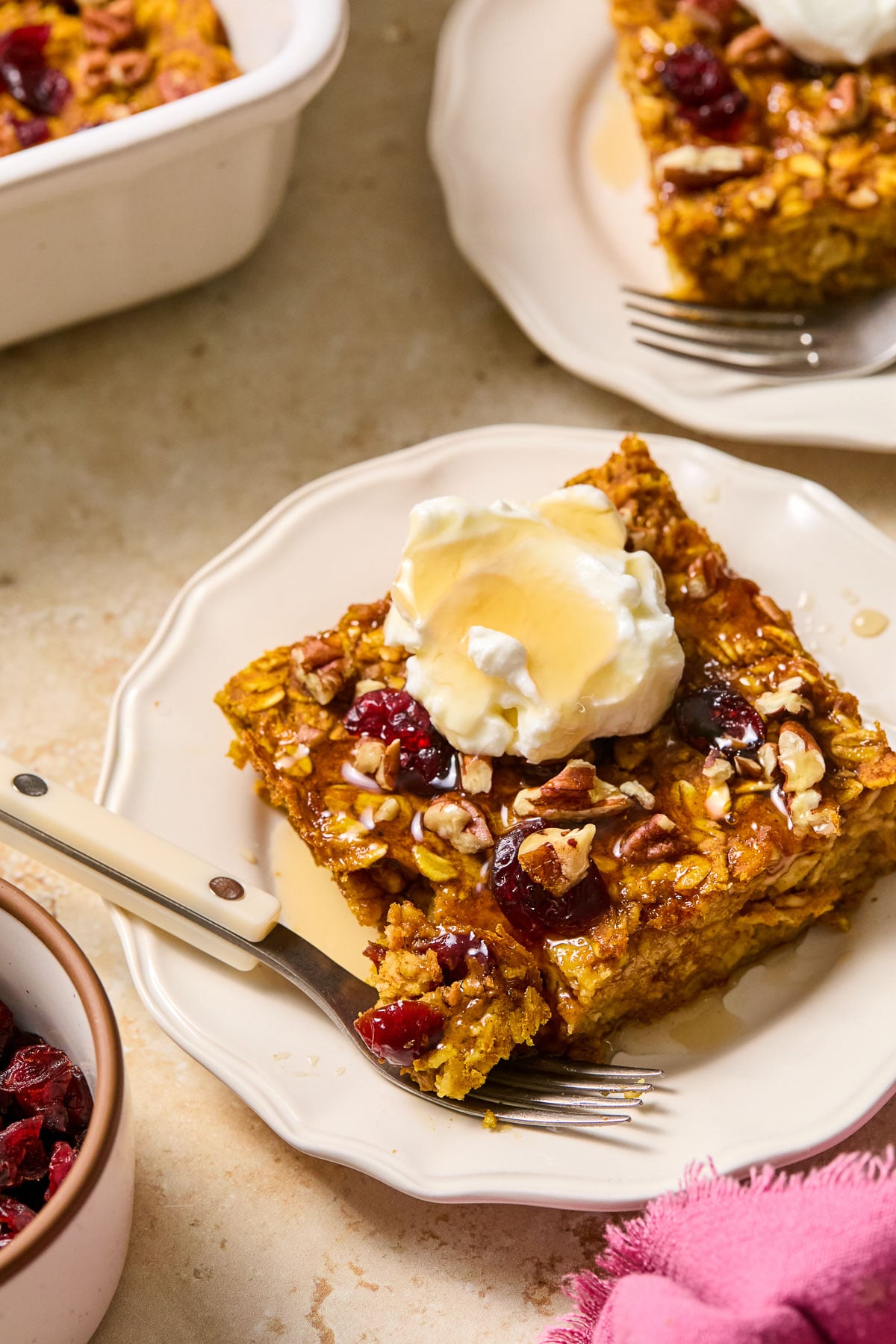 A piece of pumpkin baked oatmeal with cranberries and pecans on a white plate. It is topped with greek yogurt and a drizzle of maple syrup. A little piece is taken off the corner and is sitting on a fork on the plate. Around the plate is a pink napkin, a dish of dried cranberries, a baking dish with baked oatmeal, and another plate with a piece of baked oatmeal.