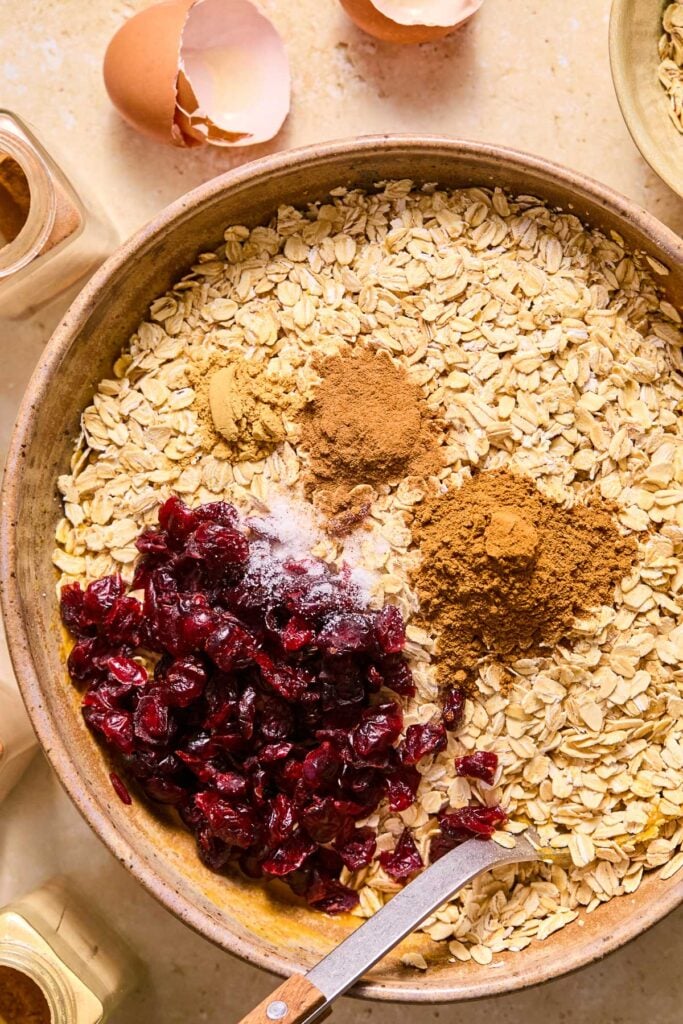 A large bowl with a wet batter topped with rolled oats, dried cranberries, and spices before it is mixed. A spoon in the bowl as well. Around the bowl are some jars of spices, eggshells, and a bowl of rolled oats.