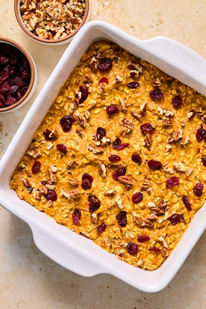A large square baking dish filled with a pumpkin oatmeal mixture topped with dried cranberries and pecans before baking.