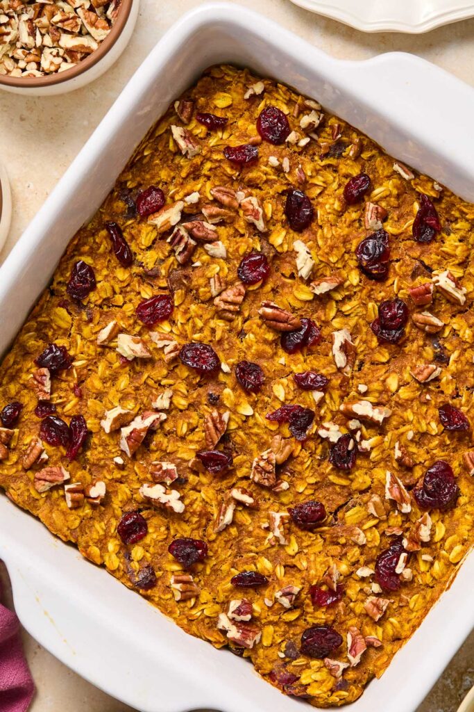 Pumpkin oatmeal bake topped with dried cranberries and pecans in a baking dish. Around the dish is a bowl of pecans.