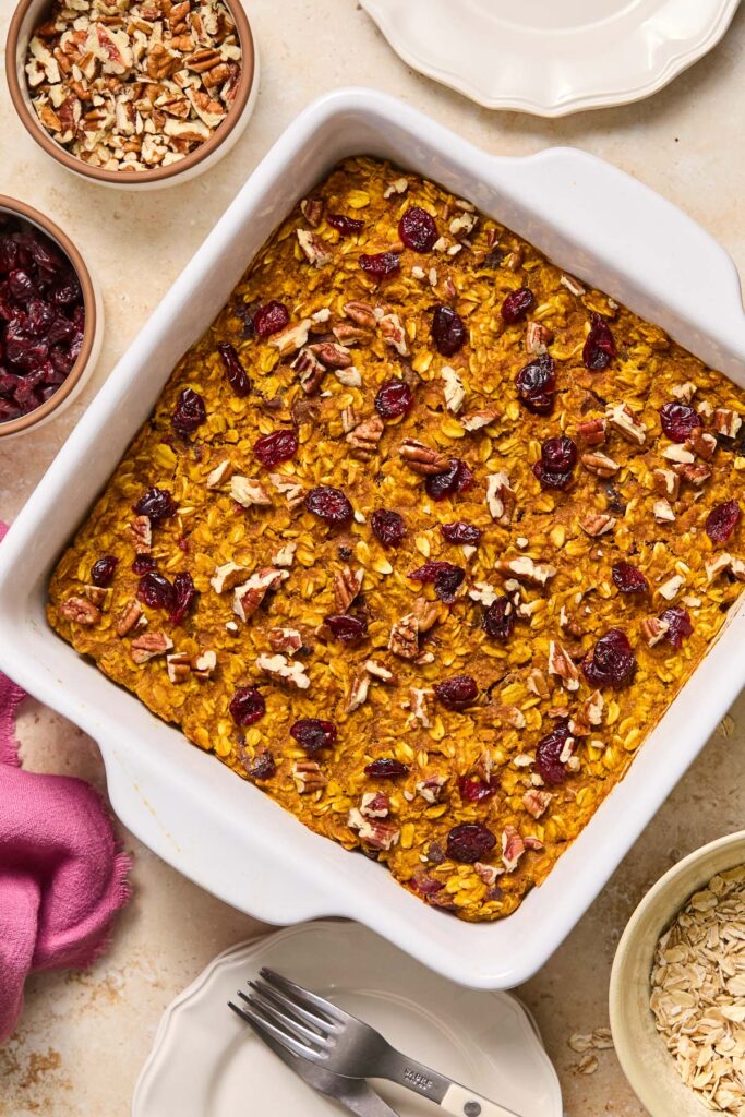 Pumpkin pie baked oatmeal topped with dried cranberries and pecans in a large baking dish. Around the dish is a bowl of rolled oats, a small plate with a fork on it, a pink napkin, a bowl of dried cranberries, a bowl of chopped pecans, and another small plate.