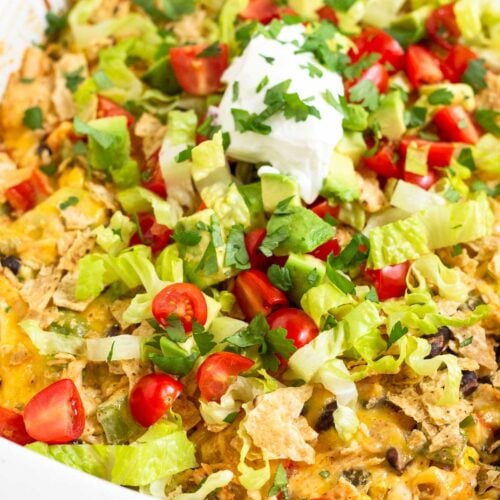 A close up of chicken taco casserole in a large baking dish topped with tortilla chips, shredded lettuce, diced tomatoes, diced avocado, sour cream, and cilantro.