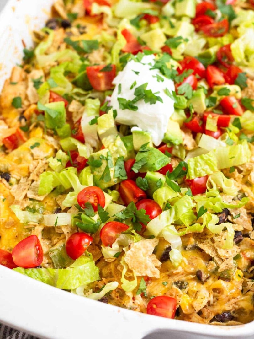 A close up of chicken taco casserole in a large baking dish topped with tortilla chips, shredded lettuce, diced tomatoes, diced avocado, sour cream, and cilantro.