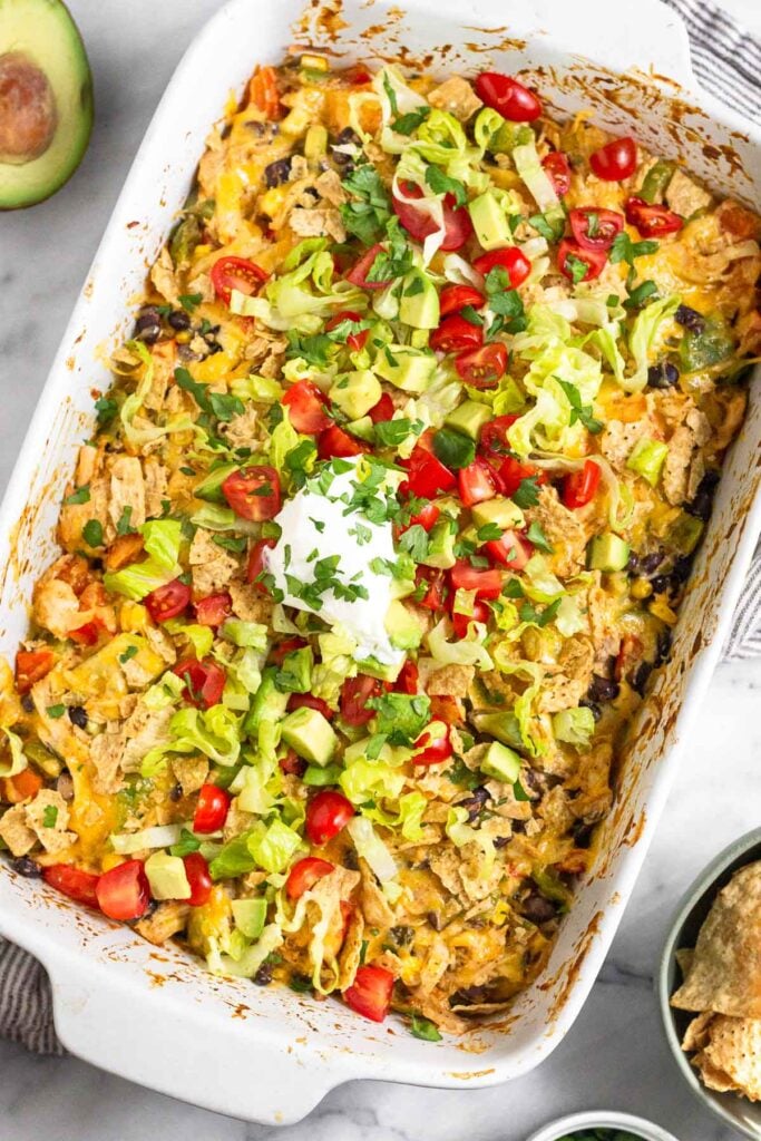 Mexican chicken taco casserole in a large baking dish. It is topped with melted cheese, tortilla chips, shredded lettuce, diced tomatoes, diced avocado, sour cream, and cilantro. Around the dish is a bowl of tortilla chips and half an avocado.