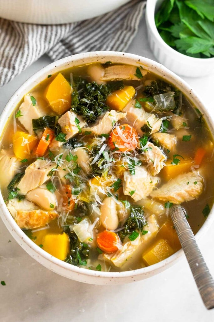 Turkey soup with butternut squash and veggies topped with herbs and cheese in a bowl. A spoon is in the bowl and next to the bowl is a ramekin of fresh herbs.
