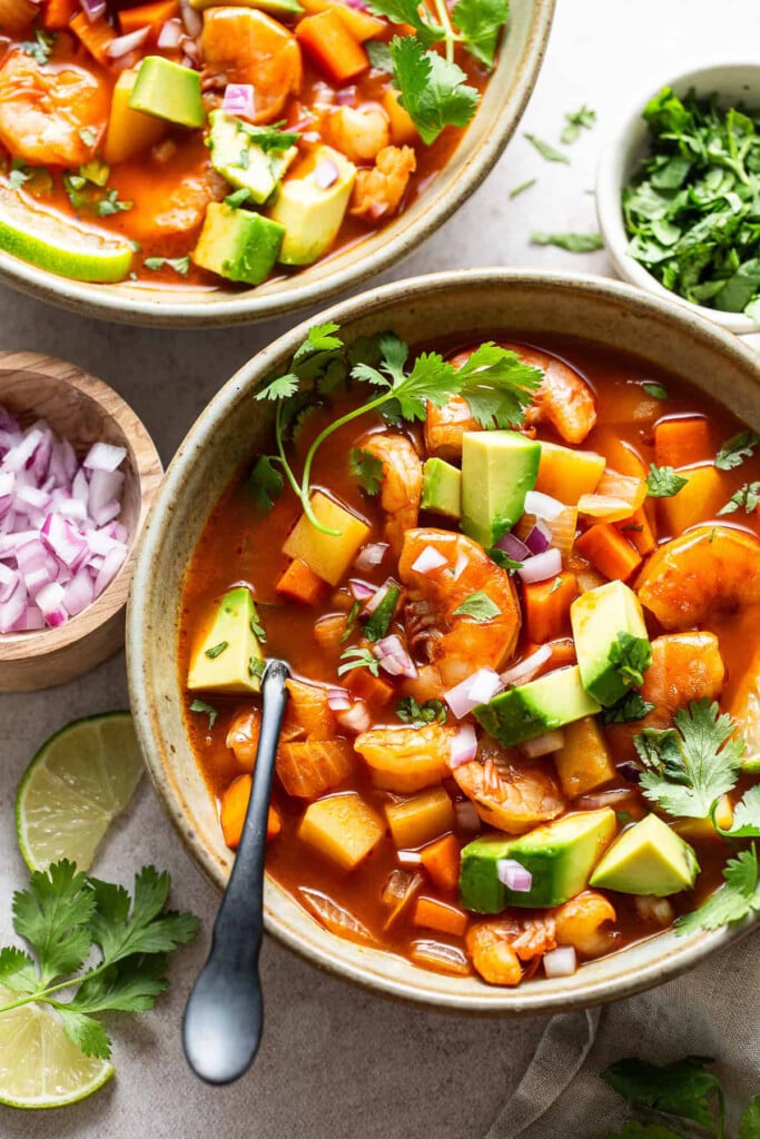 A bowl of caldo de camaron topped with diced red onion, avoacdo, and cilantro with a spoon in the bowl. Next to the bowl is another bowl of soup, a ramekin of cilantro, a ramekin of diced red onion, and a couple of lime wedges.