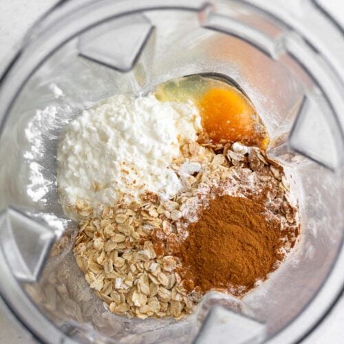 A blender filled with cottage cheese, eggs, oats, cinnamon, and baking powder before it is blended together.